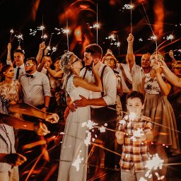 Janusz i Agata - Para młoda w objęciach, otoczona gośćmi weselnymi z zimnymi ogniami, w tle ciemne niebo, oświetlenie ciepłe, tworzące efektowne smugi świetlne.