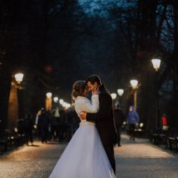 Janusz i Agata - Para młoda obejmuje się na oświetlonej alei parkowej nocą, panna młoda w białej sukni i futrzanym bolerku, pan młody w ciemnym płaszczu, w tle rozmyte sylwetki przechodniów i rzędy parkowych lamp
