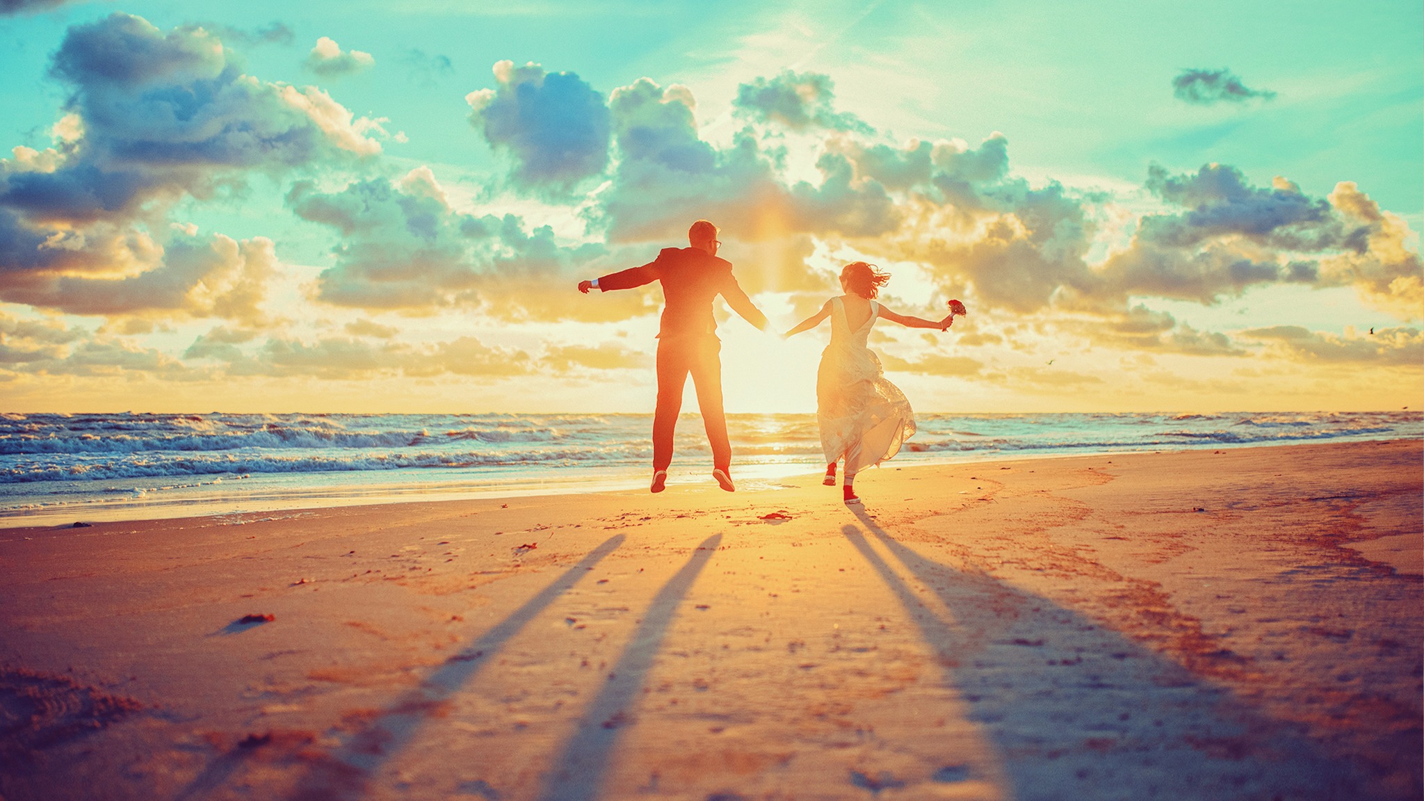 Para młoda skacze na plaży o zachodzie słońca, trzymając się za ręce, z długimi cieniami rzucanymi na piasek.