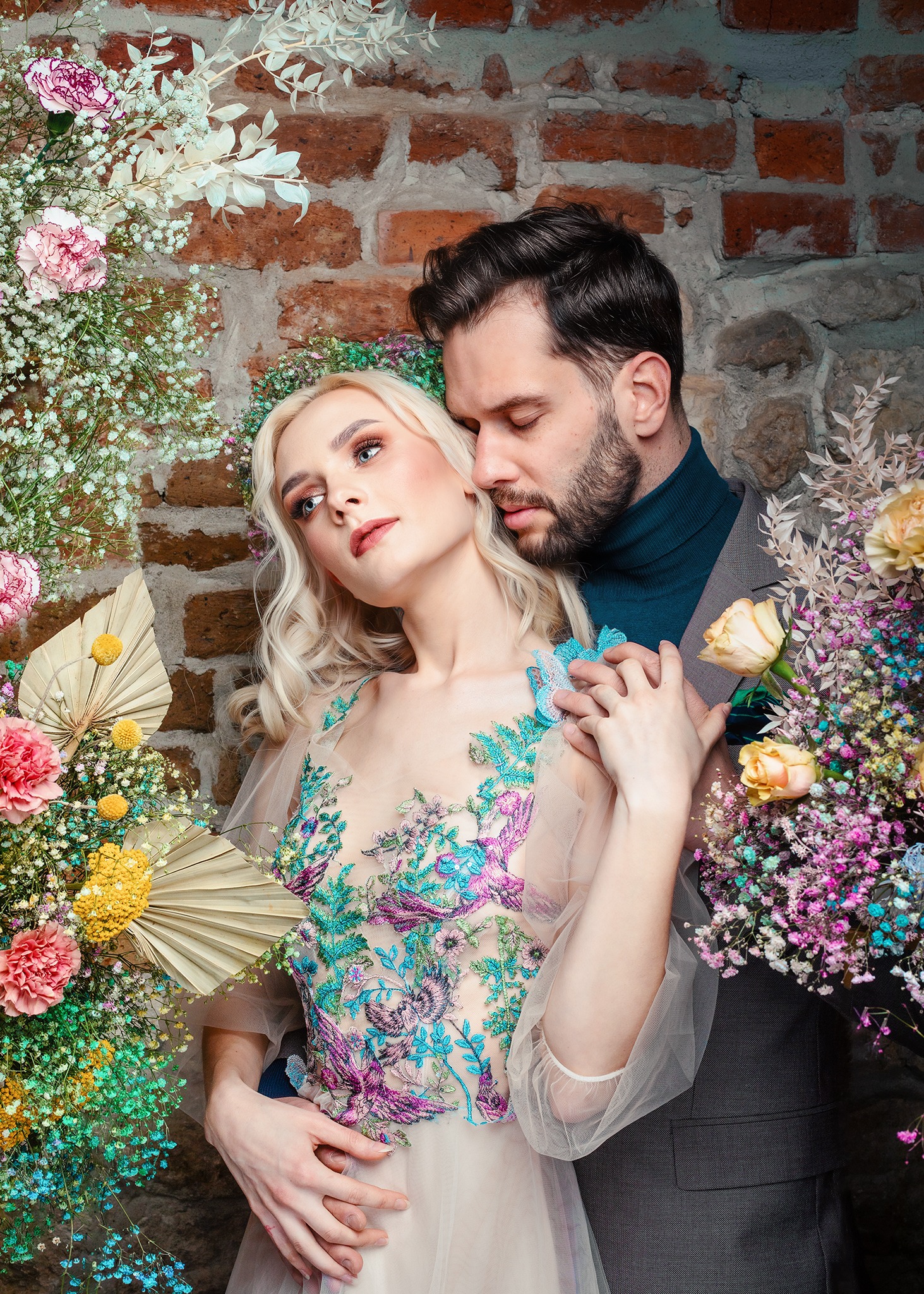 Para w otoczeniu kwiatów na tle ceglanej ściany: kobieta w sukni z haftem kwiatowym i wianku, mężczyzna w garniturze i golfie.