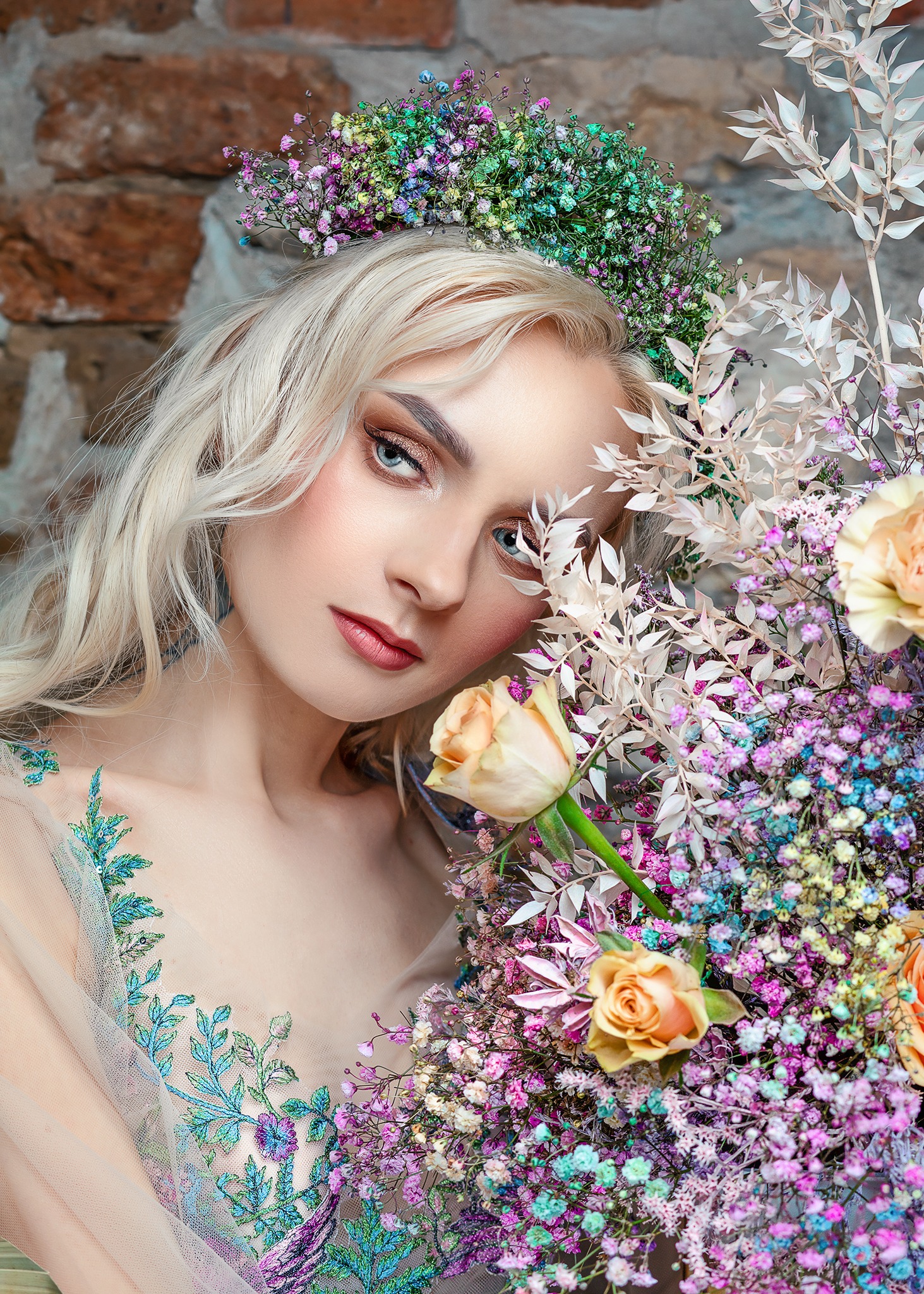 Portret kobiety o blond włosach i niebieskich oczach, z wiankiem z kolorowych kwiatów na głowie, pozującej z bukietem róż i gipsówki na tle ceglanej ściany.
