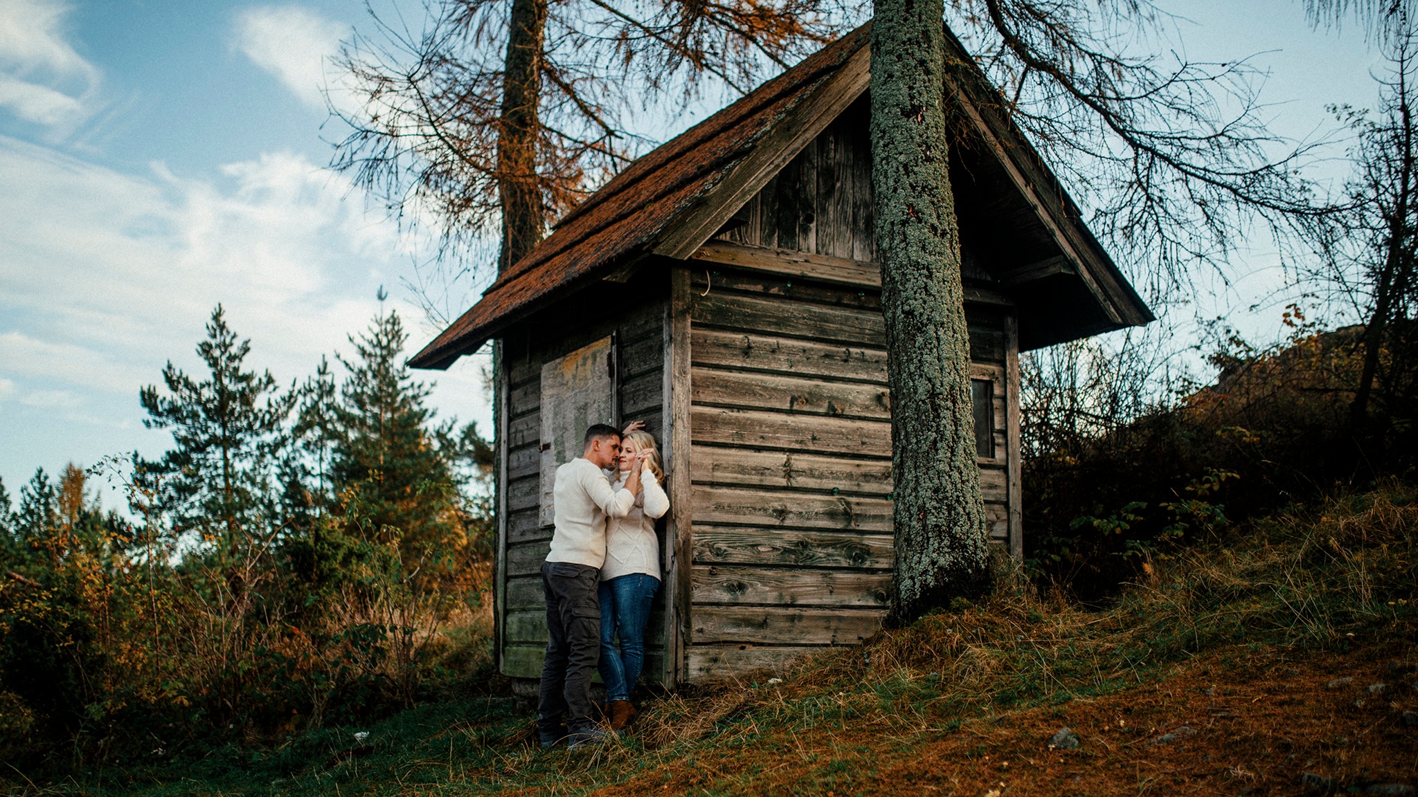 Zakochana para w białych swetrach przytulona przy drewnianej chatce z brązowym dachem, otoczona drzewami i trawą w jesiennej scenerii.