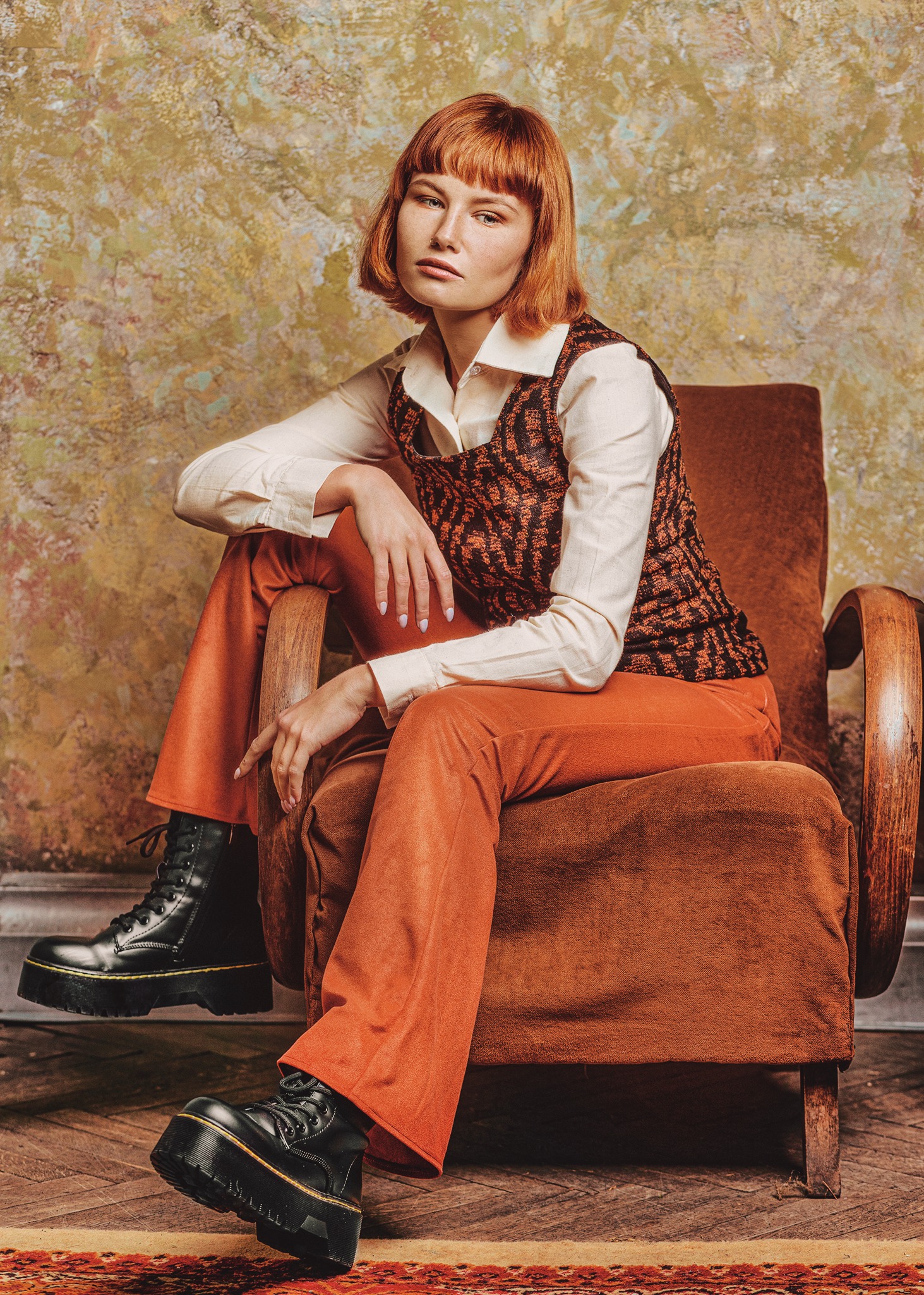 Kobieta z rudymi włosami w stylu lat 70. siedzi na brązowym fotelu w pomarańczowych spodniach i czarnych botkach na platformie, pozując na tle teksturowanej ściany.