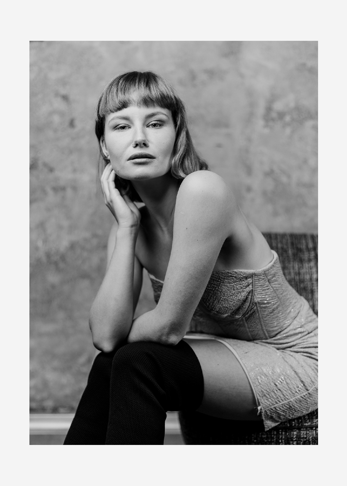 Czarno-biały portret kobiety z krótką grzywką, w sukience z cekinami i wysokich czarnych podkolanówkach, opierającej brodę na dłoni, z kolanami podpartymi rękami na tle teksturowanej ściany.