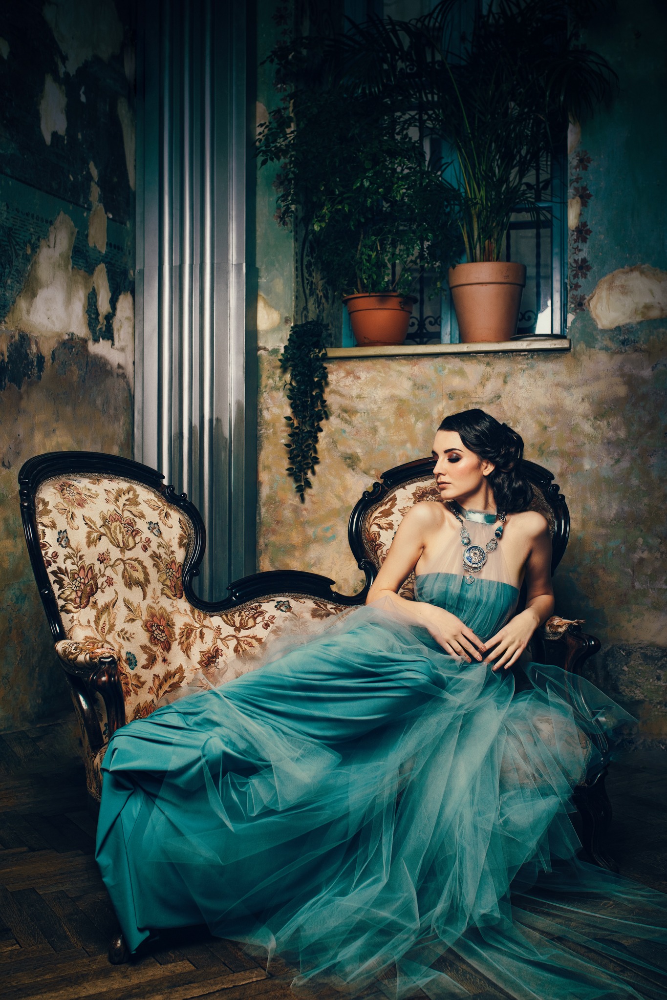 Kobieta w turkusowej sukni balowej siedzi na zdobionym fotelu w pokoju z roślinami doniczkowymi i ścianami w stylu vintage.