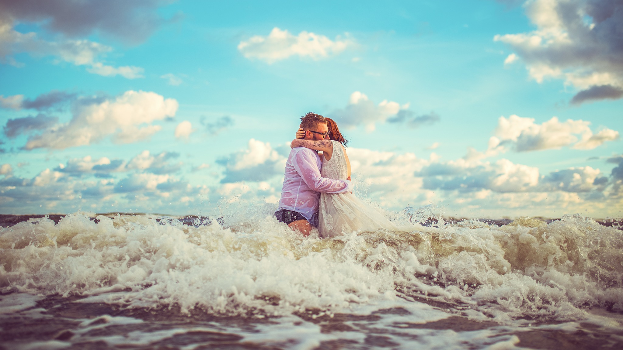 Para młoda w objęciach w falach morskich; panna młoda w białej sukni, pan młody w mokrej, białej koszuli i czarnych spodenkach, niebo z chmurami w tle.