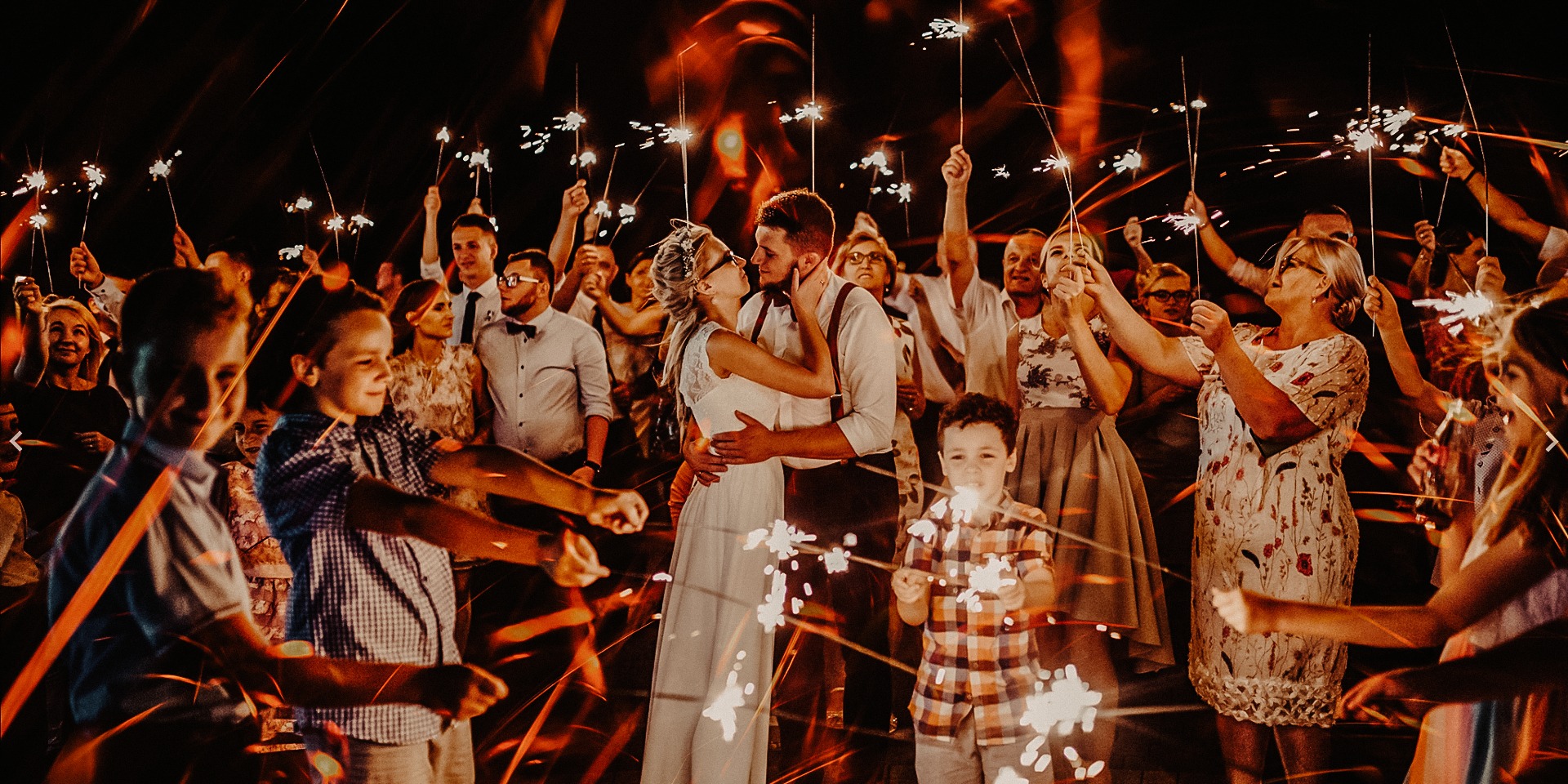 Para młoda w objęciach, otoczona gośćmi weselnymi z zimnymi ogniami, w tle ciemne niebo, oświetlenie ciepłe, tworzące efektowne smugi świetlne.