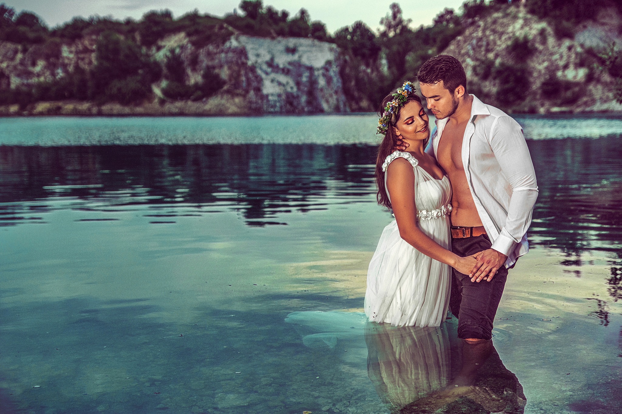 Zakochana para w białej sukni i rozpiętej koszuli, zanurzona w wodzie jeziora na tle skalistego brzegu, sesja narzeczeńska.