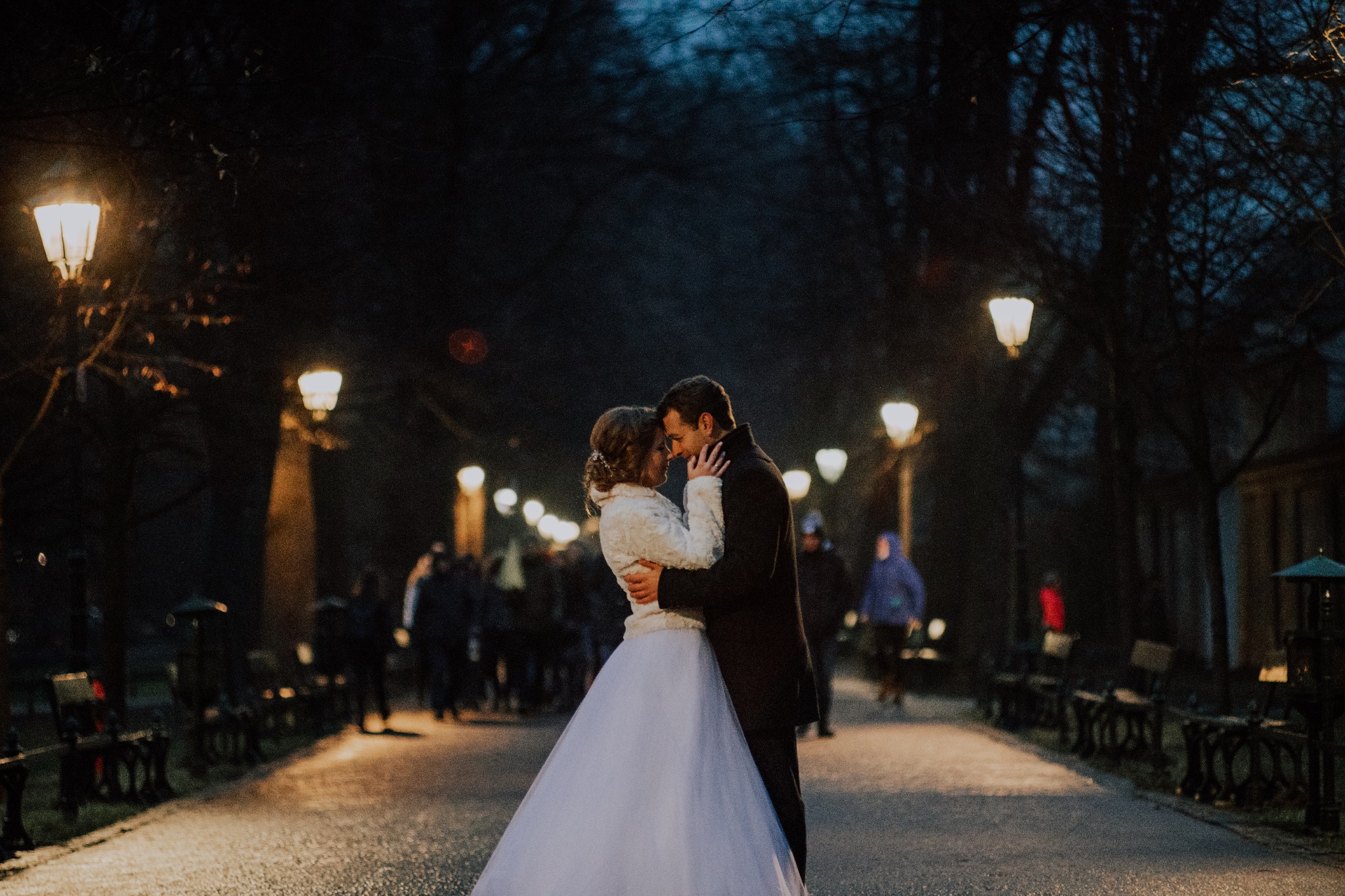 Para młoda obejmuje się na oświetlonej alei parkowej nocą, panna młoda w białej sukni i futrzanym bolerku, pan młody w ciemnym płaszczu, w tle rozmyte sylwetki przechodniów i rzędy parkowych lamp