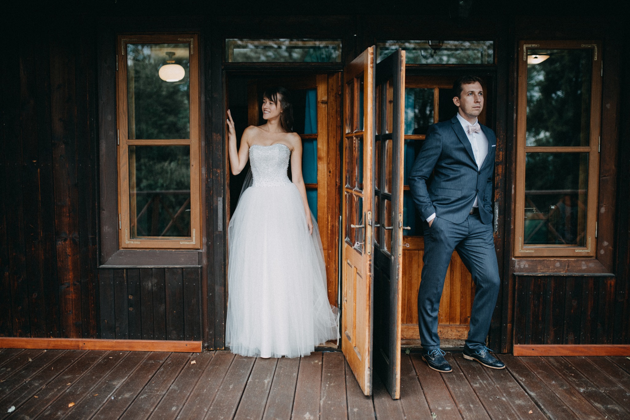 Para młoda pozuje w otwartych drzwiach drewnianego domu; panna młoda w białej sukni bez ramiączek, pan młody w szarym garniturze i różowej muszce.