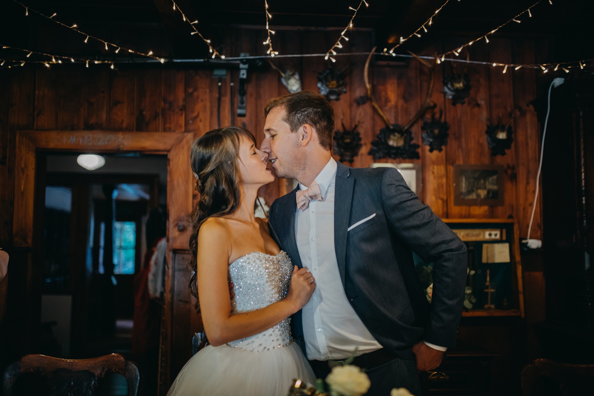 Pocałunek pary młodej w rustykalnym wnętrzu z drewnianymi ścianami, porożami i dekoracyjnymi lampkami.