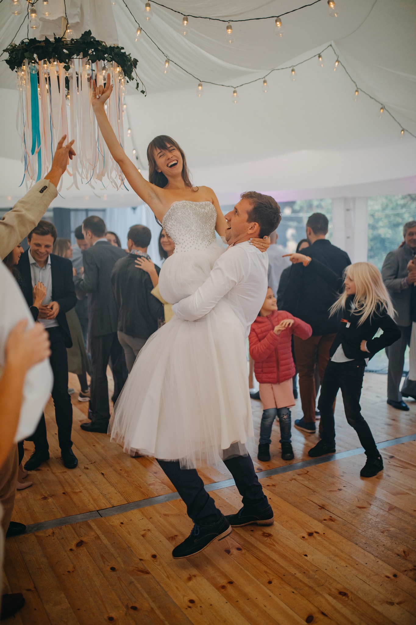 Uśmiechnięty pan młody podnosi pannę młodą w sukni bez ramiączek podczas weselnego przyjęcia pod namiotem, w tle goście i drewniany parkiet