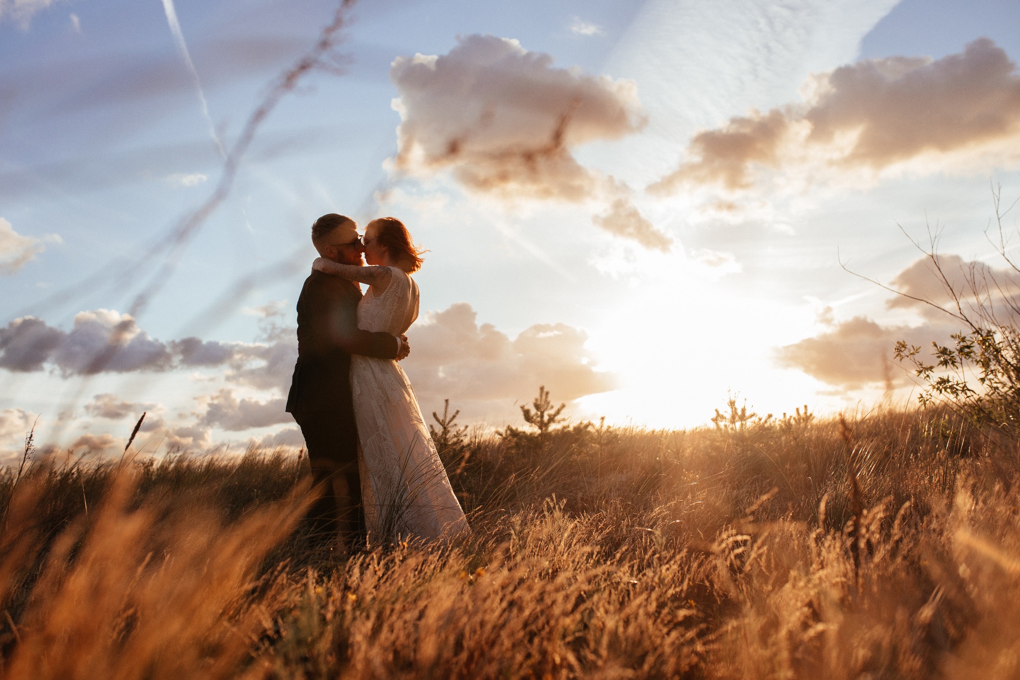Para młoda w objęciach na tle zachodzącego słońca i wysokiej trawy, mężczyzna w ciemnym garniturze i okularach, kobieta w koronkowej sukni z odkrytymi plecami.
