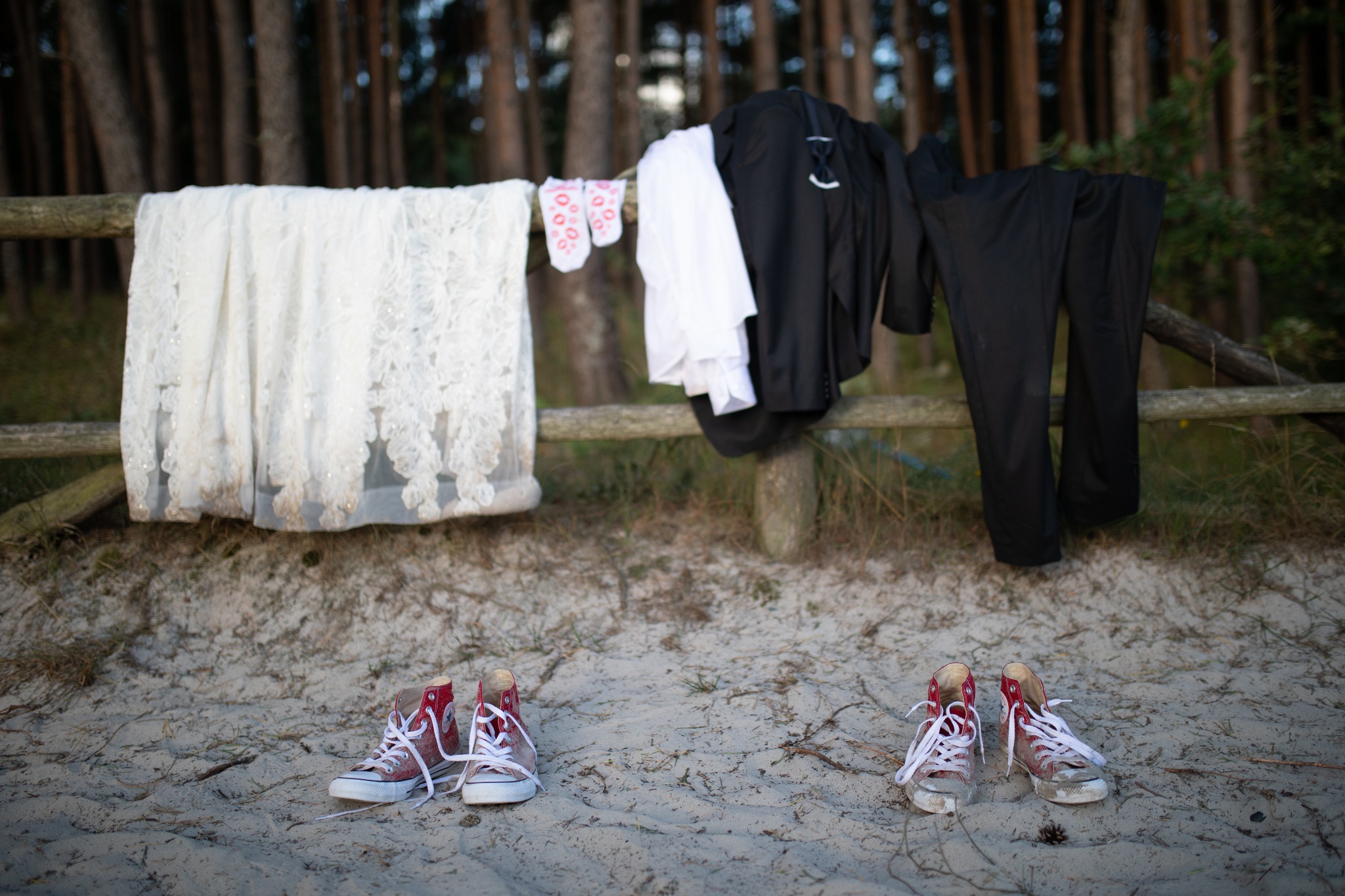Ślubna suknia z koronką, męski garnitur, skarpetki w serduszka i para czerwonych trampek leżą na piasku przed drewnianym płotem w lesie.