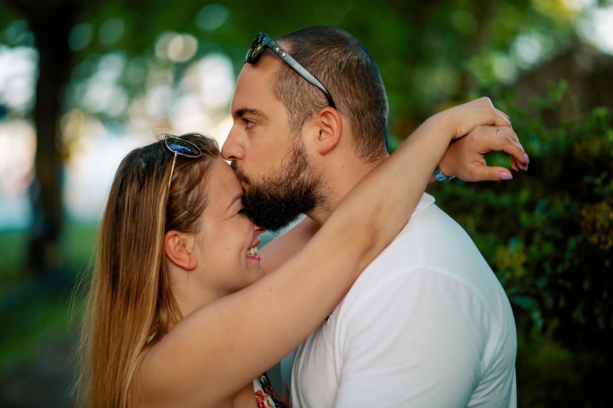 Mężczyzna w okularach całuje uśmiechniętą kobietę w czoło, obejmując ją ramionami na tle zieleni.