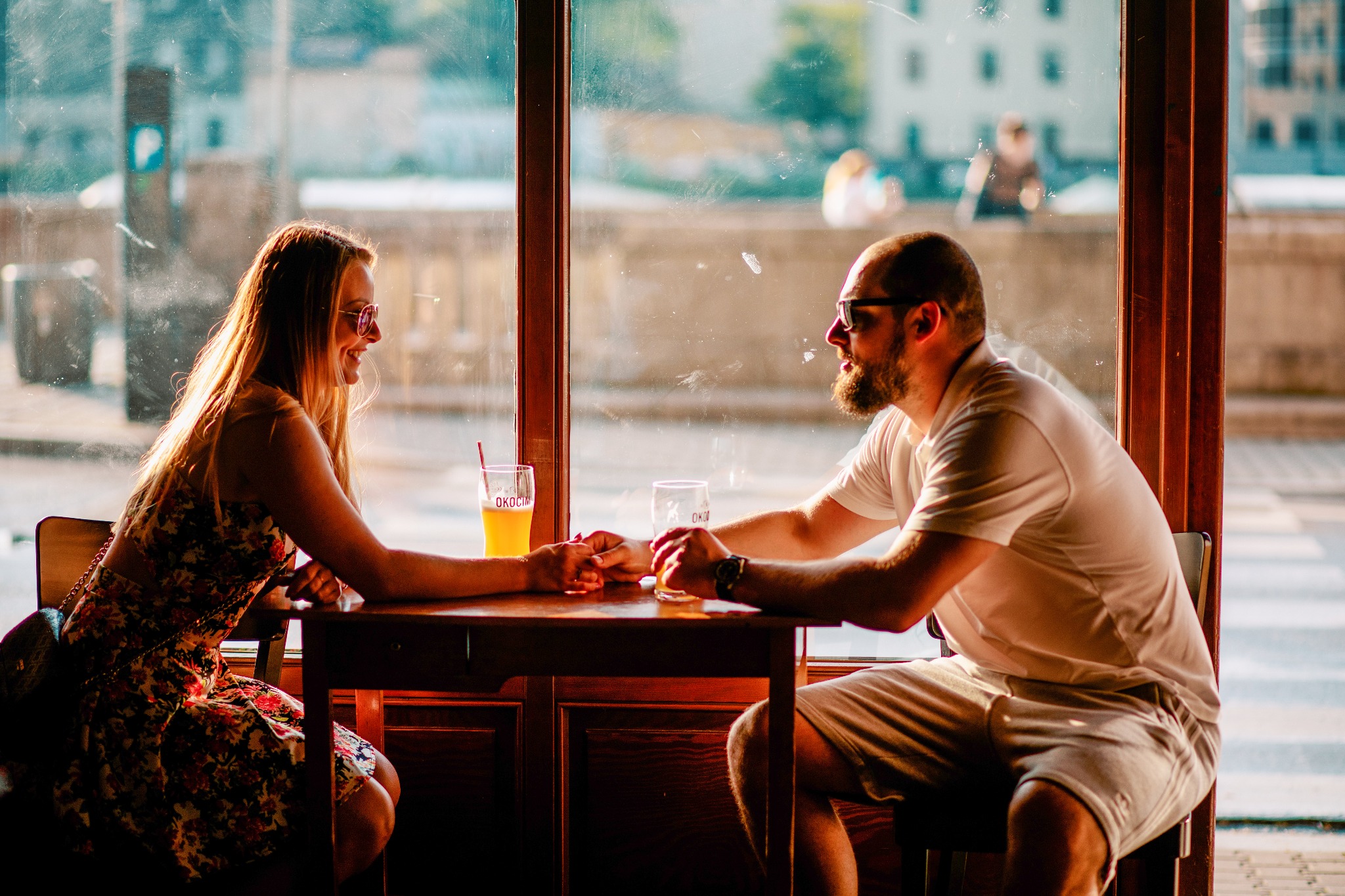 Para siedząca przy stoliku w kawiarni, trzymająca się za ręce, z napojami i widokiem na miasto w tle.