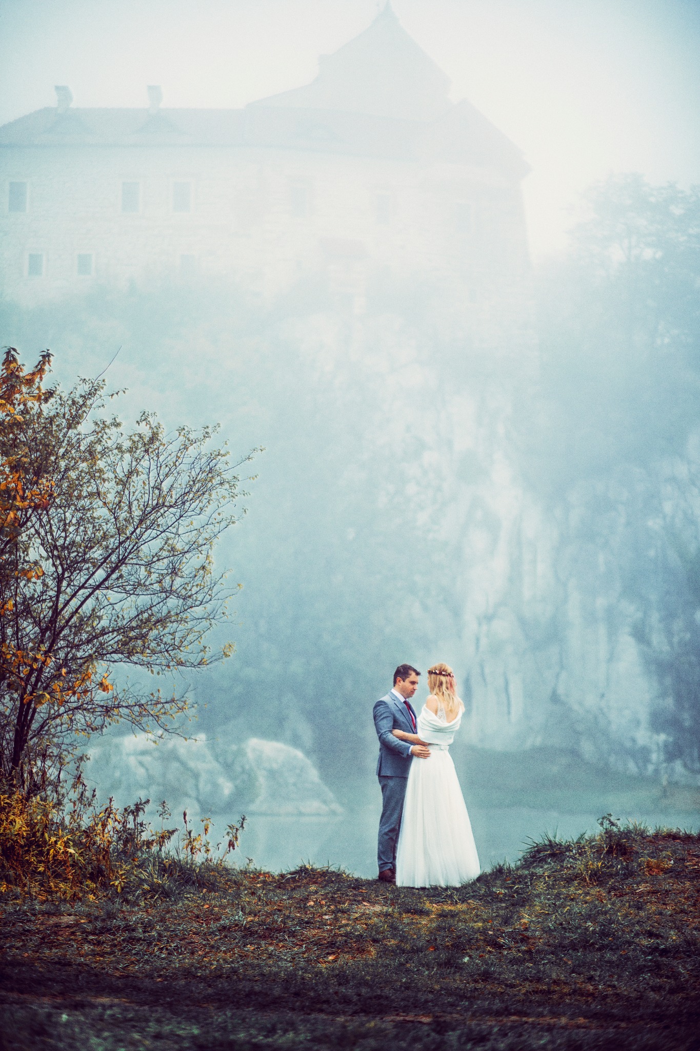 Para młoda w objęciach na wzgórzu, z zamkiem spowitym mgłą w tle, jesienne drzewo po lewej stronie kadru, romantyczna atmosfera.