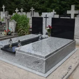 Podwójny, jasnoszary pomnik z czarnymi tablicami, z krzyżem pośrodku, kwiatami i lampionami, na cmentarzu.