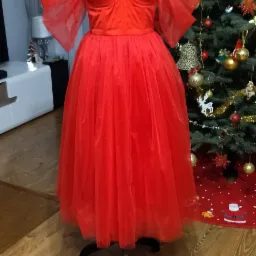 Czerwona, tiulowa suknia z odkrytymi ramionami na manekinie krawieckim, z choinką w tle.