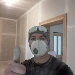 Mężczyzna w masce i okularach ochronnych, z uniesionym kciukiem do góry, stoi w pomieszczeniu z niedokończonymi ścianami z płyt kartonowo-gipsowych, widoczne łączenia płyt.