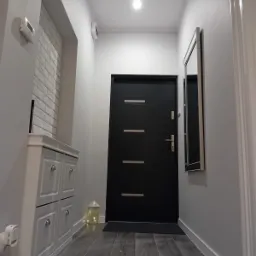 Nowoczesny przedpokój w szarościach z czarnymi drzwiami wejściowymi, drewnopodobna podłoga, biała cegła na ścianie i szafka na buty z czterema szufladami.