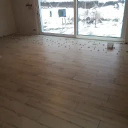 Wnętrze w trakcie układania podłogi z paneli drewnopodobnych; widoczne dystanse, okno z widokiem na zaśnieżony ogród i fragment kominka.