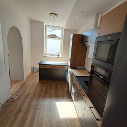 FIRMA USŁUGOWA ADRIAN KALINOWSKI - Nowoczesna kuchnia z drewnianą podłogą i zabudową w odcieniach szarości oraz drewna. Widoczny sprzęt AGD marki Amica. W tle okno z wiszącą lampą.