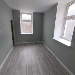 FIRMA USŁUGOWA ADRIAN KALINOWSKI - Pomalowane na szaro wnętrze pokoju z panelami podłogowymi i dwoma oknami. Widok na ściany, podłogę i okna. Świeżo po remoncie.