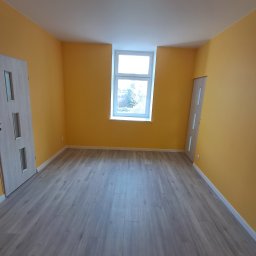 FIRMA USŁUGOWA ADRIAN KALINOWSKI - Pokój po remoncie: świeżo pomalowane ściany na żółto, panele podłogowe w odcieniach szarości, białe drzwi z przeszkleniami i okno z widokiem na zieleń.