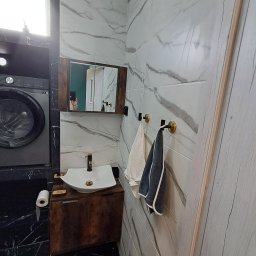 FIRMA USŁUGOWA ADRIAN KALINOWSKI - Wykończona łazienka z marmurowymi płytkami, umywalką na drewnianej szafce, pralką i lustrem. Widoczne ręczniki na złotych haczykach i rolka papieru toaletowego.