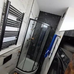 Nowoczesna łazienka z czarną kabiną prysznicową, grzejnikiem drabinkowym i pralką. Marmurowe płytki na ścianach i podłodze. Elementy wykończenia w kolorze złotym.