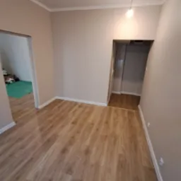 Jasne panele podłogowe w korytarzu po remoncie, widoczne listwy przypodłogowe i wykończenie ścian w neutralnym odcieniu. W tle widoczne otwarte drzwi do innych pomieszczeń.