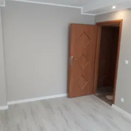 Pomalowane na szaro ściany w pokoju z panelami podłogowymi i otwartymi drewnianymi drzwiami z romboidalnymi zdobieniami.