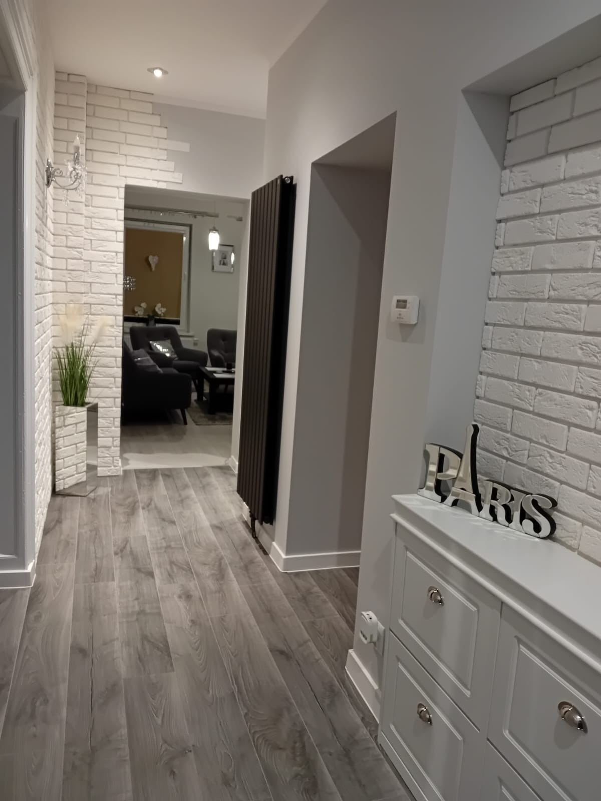 Nowoczesny korytarz w Wałbrzychu z szarą podłogą imitującą drewno, białą cegłą na ścianach, grzejnikiem panelowym i białym kredensem z dekoracją 'Paris'.