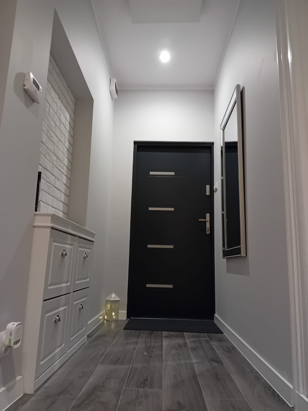 Nowoczesny przedpokój w szarościach z czarnymi drzwiami wejściowymi, drewnopodobna podłoga, biała cegła na ścianie i szafka na buty z czterema szufladami.