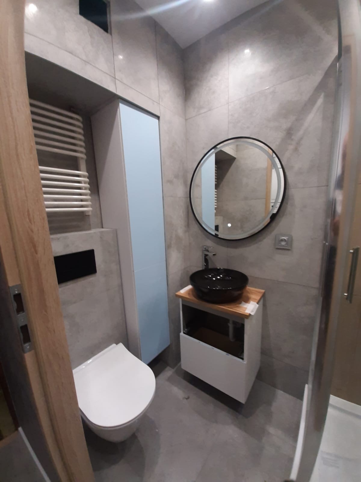 Nowoczesna łazienka z szarymi płytkami, okrągłym lustrem z podświetleniem LED, czarną umywalką nablatową na drewnianym blacie, wiszącą toaletą i grzejnikiem drabinkowym.