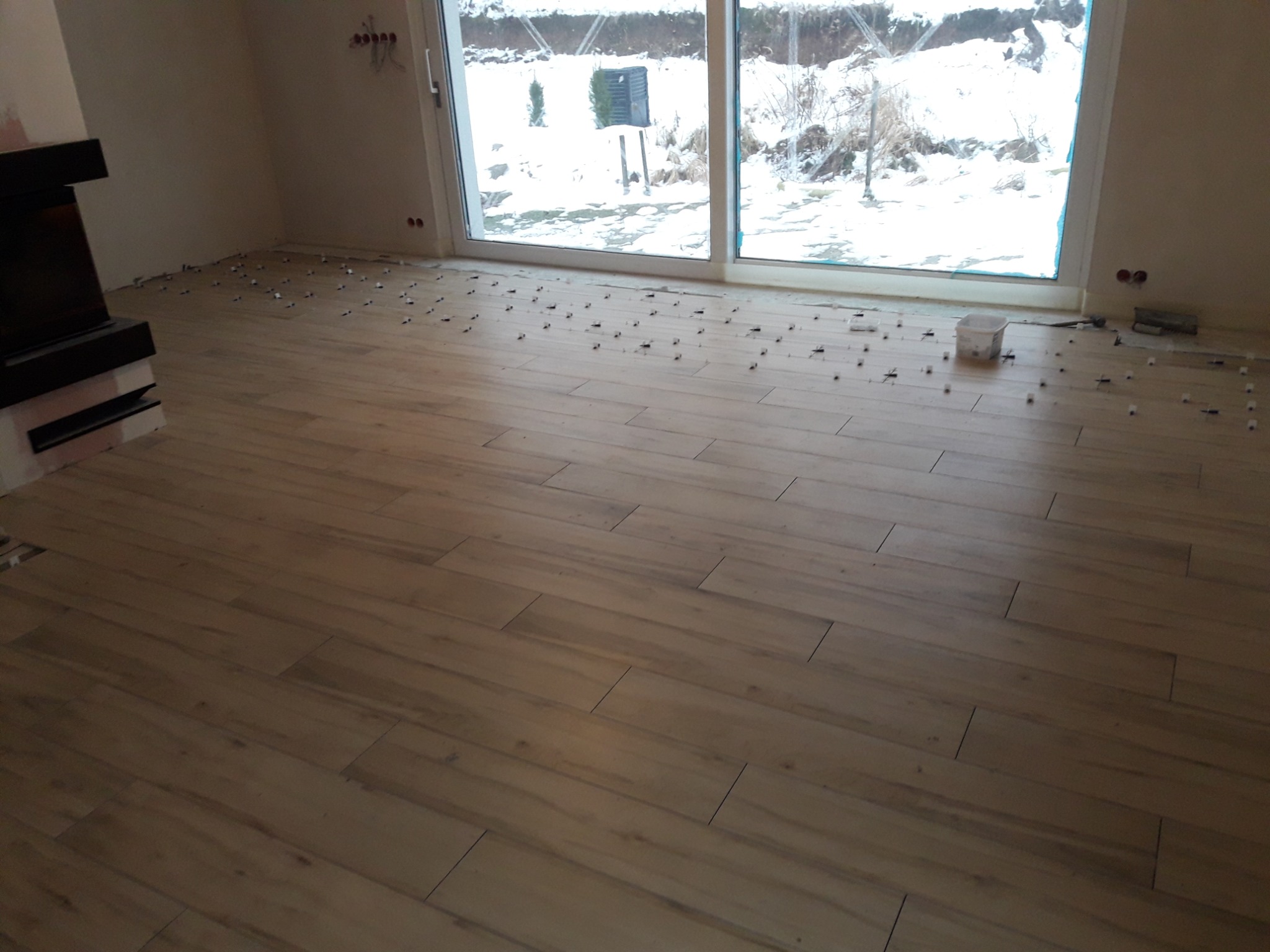 Wnętrze w trakcie układania podłogi z paneli drewnopodobnych; widoczne dystanse, okno z widokiem na zaśnieżony ogród i fragment kominka.