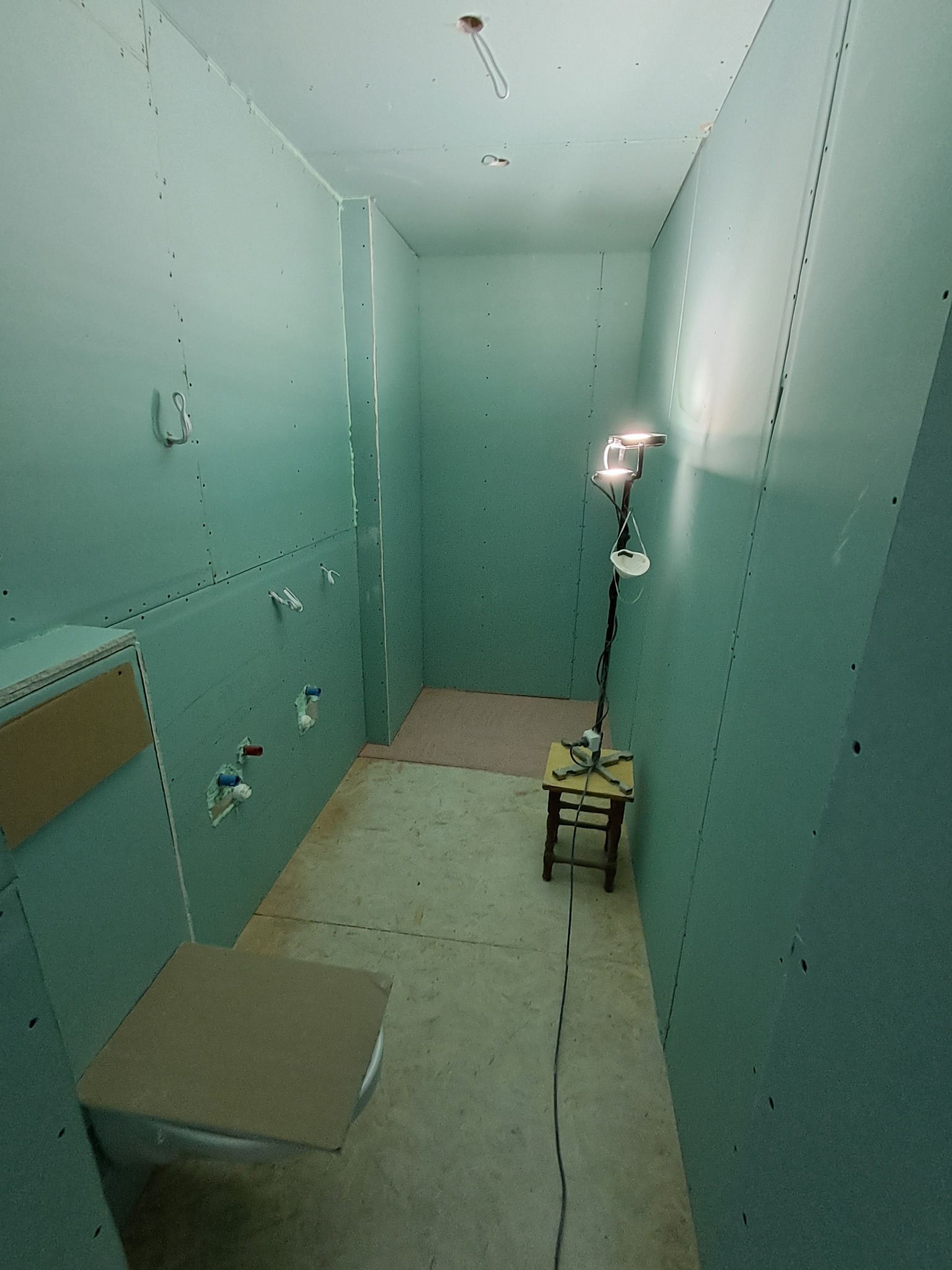 Wnętrze łazienki w trakcie prac wykończeniowych: ściany obłożone płytami karton-gips, widoczne instalacje hydrauliczne, prowizoryczne oświetlenie i narzędzia.