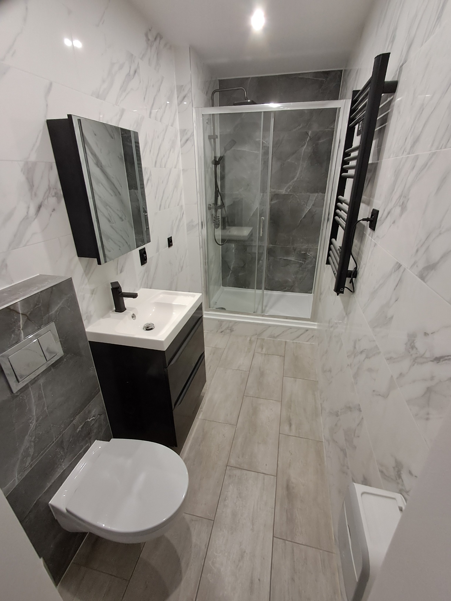 Nowoczesna łazienka po remoncie: białe marmurowe płytki, czarna armatura, kabina prysznicowa z szarymi płytkami i grzejnik drabinkowy. Minimalistyczny design.