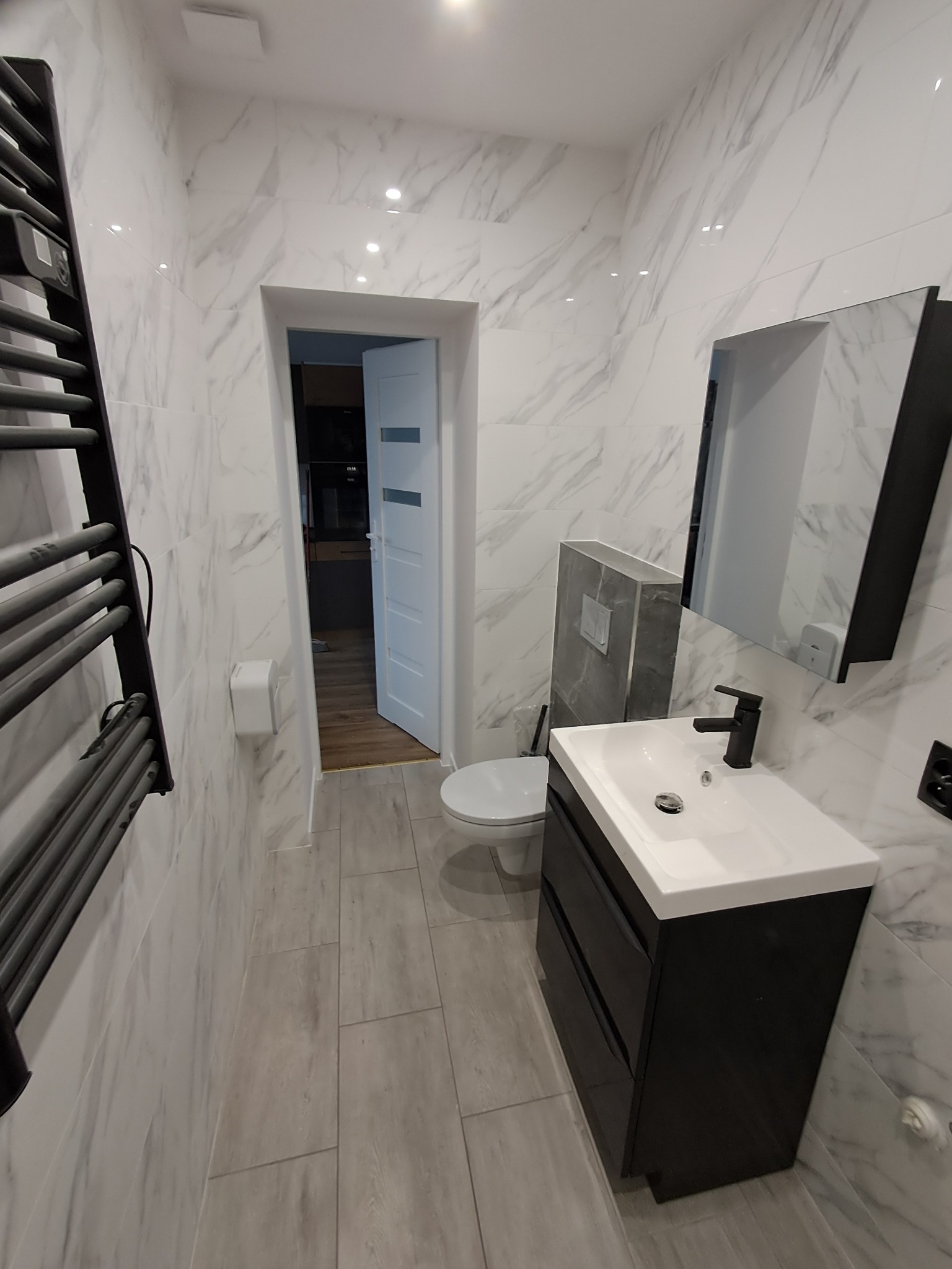 Biało-szara łazienka z marmurowymi płytkami, podwieszaną toaletą i czarną szafką umywalkową. Widoczny grzejnik drabinkowy i otwarte drzwi do innego pomieszczenia.