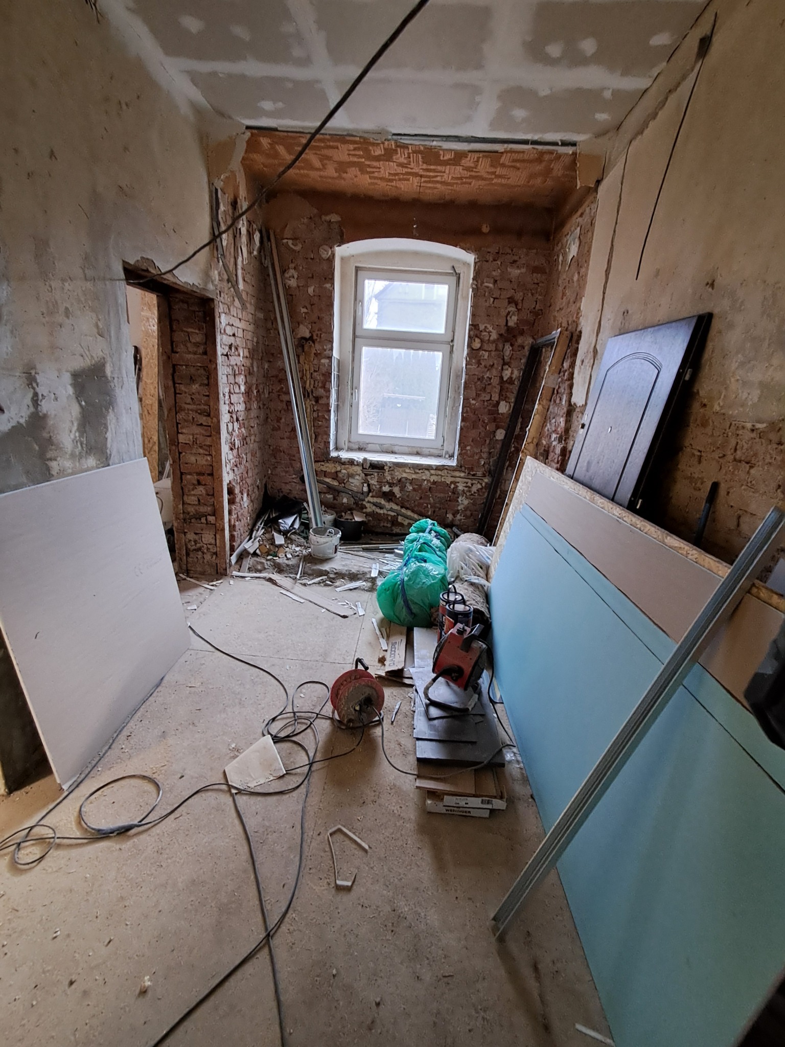 Wnętrze w trakcie remontu: odsłonięta cegła, płyty gipsowe, narzędzia. Widoczne instalacje na suficie i okno w tle. Prace przygotowawcze przed wykończeniem.