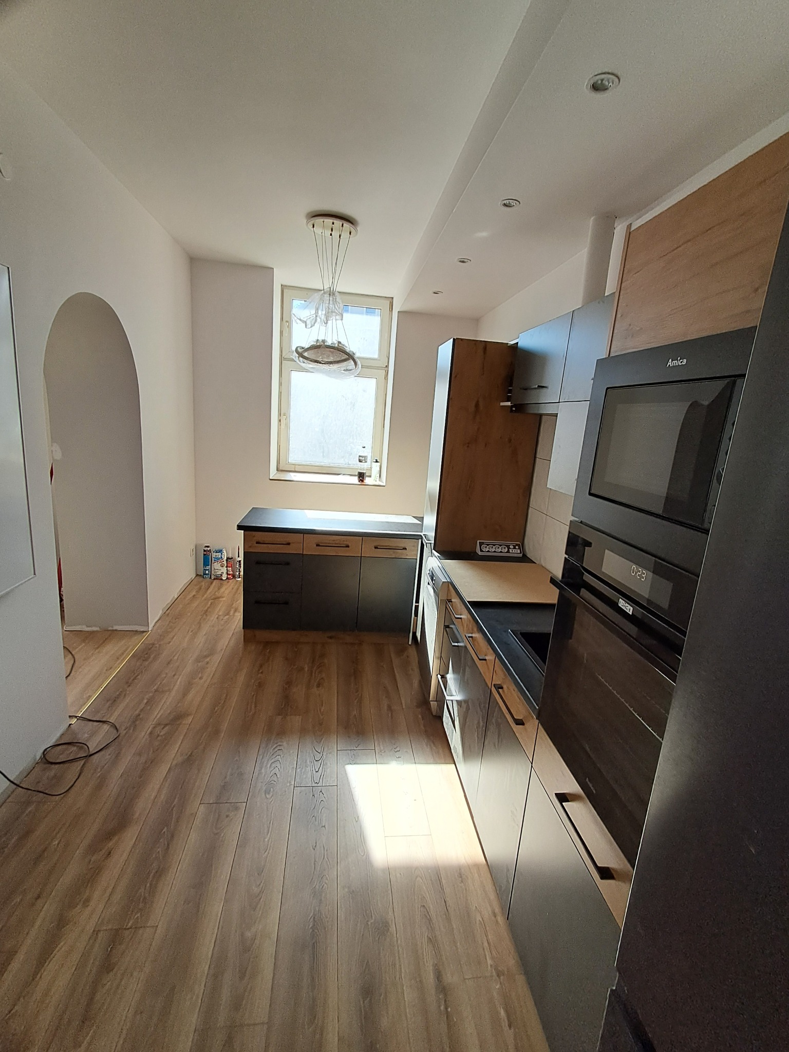 Nowoczesna kuchnia z drewnianą podłogą i zabudową w odcieniach szarości oraz drewna. Widoczny sprzęt AGD marki Amica. W tle okno z wiszącą lampą.