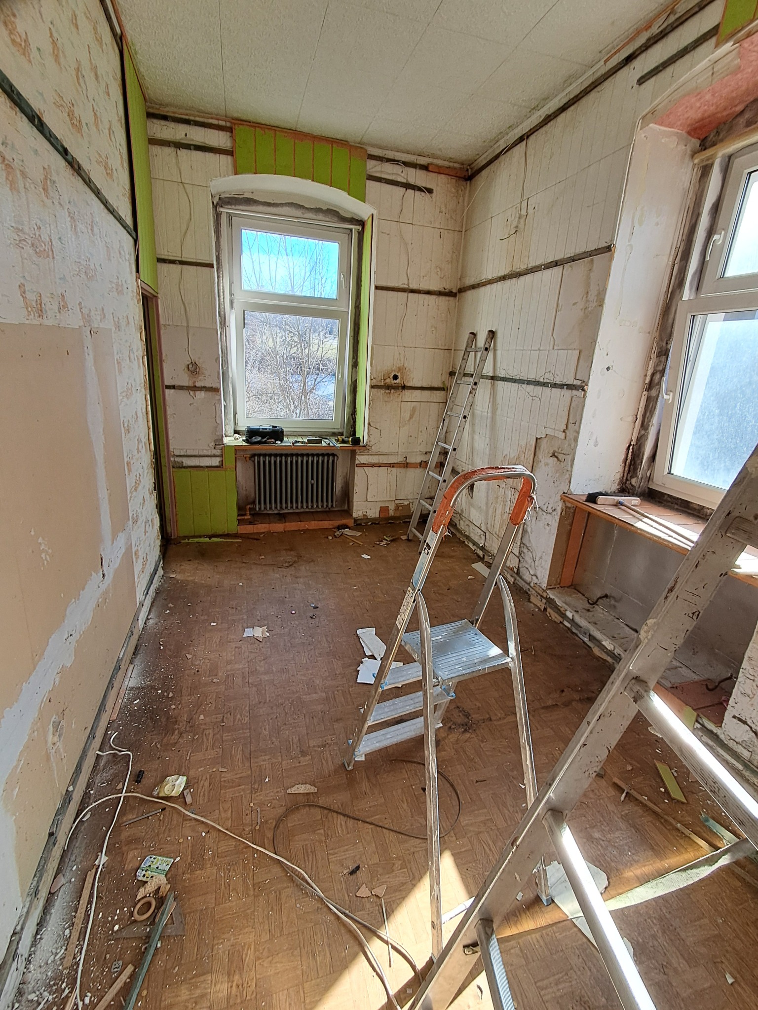 Pokój w trakcie demontażu: odsłonięte ściany, fragmenty tapet, dwie drabiny, stare okno. Widoczne ślady po instalacjach i elementy wykończeniowe na podłodze.