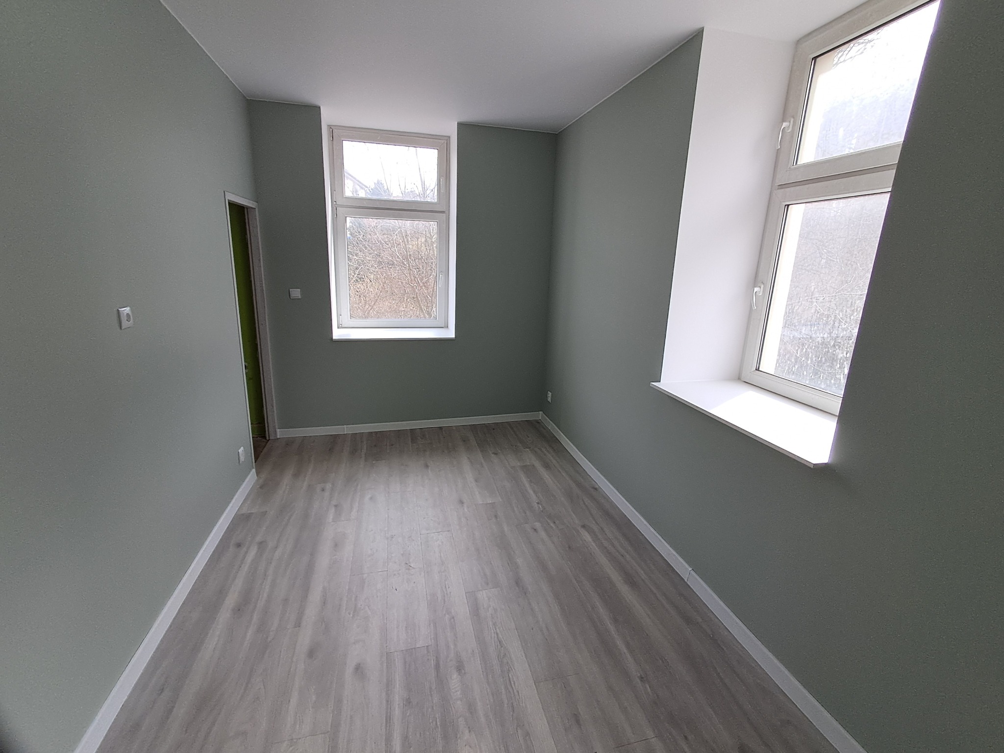 Pomalowane na szaro wnętrze pokoju z panelami podłogowymi i dwoma oknami. Widok na ściany, podłogę i okna. Świeżo po remoncie.