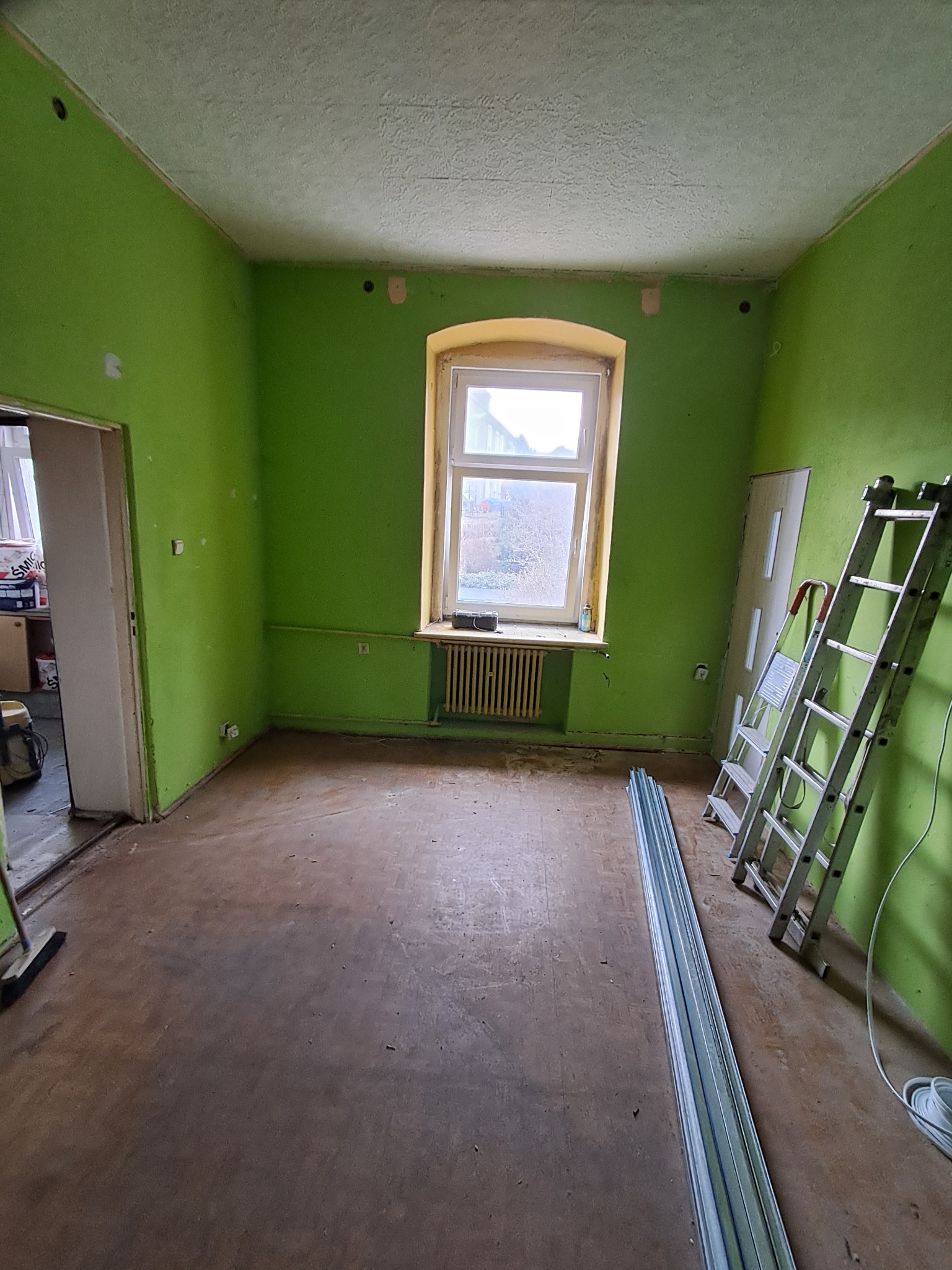 Pokój przed remontem: zielone ściany, okno z grzejnikiem, drabina oparta o ścianę, profile do płyt g-k. Widoczne ślady po instalacjach elektrycznych.