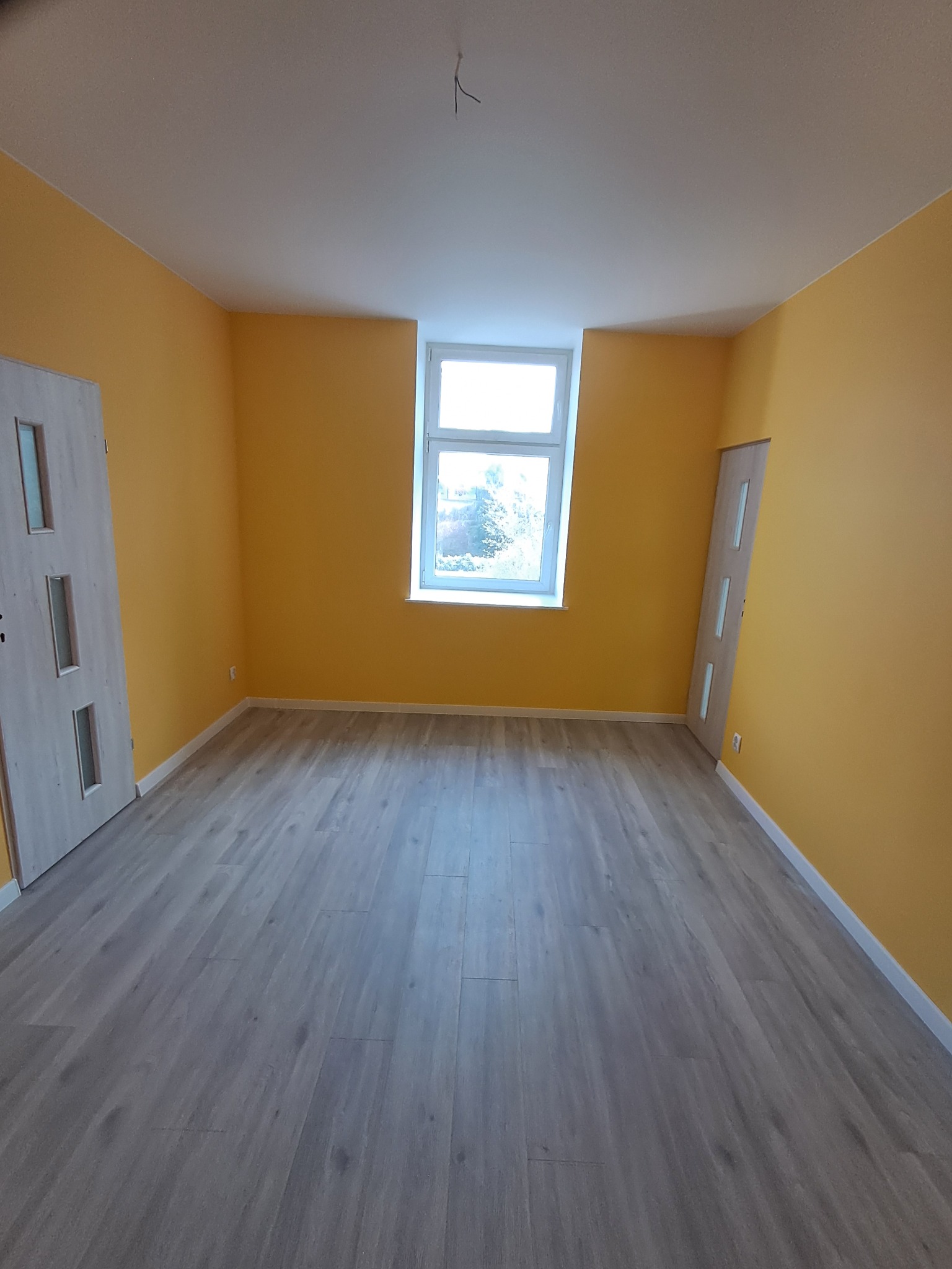 Pokój po remoncie: świeżo pomalowane ściany na żółto, panele podłogowe w odcieniach szarości, białe drzwi z przeszkleniami i okno z widokiem na zieleń.