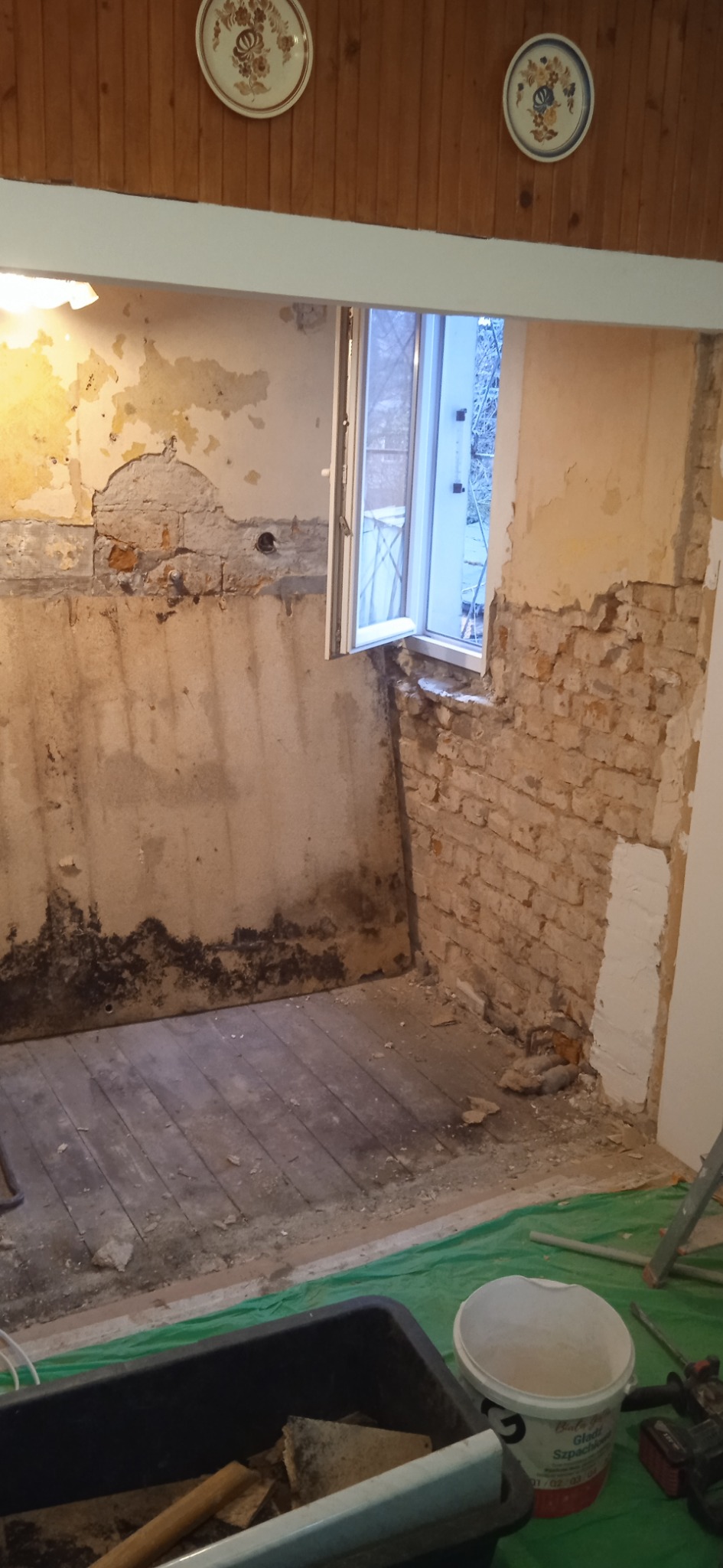 Pokój w trakcie remontu: zdarte tapety, odsłonięta cegła, widoczne ślady wilgoci. Nowe okno, drewniana podłoga częściowo zakryta folią, narzędzia w wiadrze.