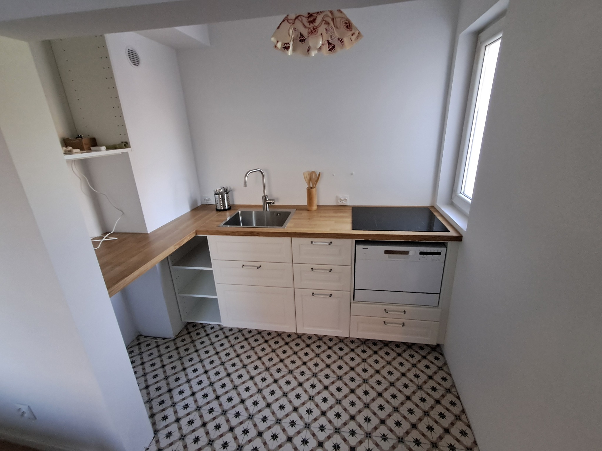 Jasna kuchnia w bloku: białe szafki z drewnianym blatem, mozaikowa podłoga, okno w skosie. Nowoczesna aranżacja z okapem i płytą indukcyjną.