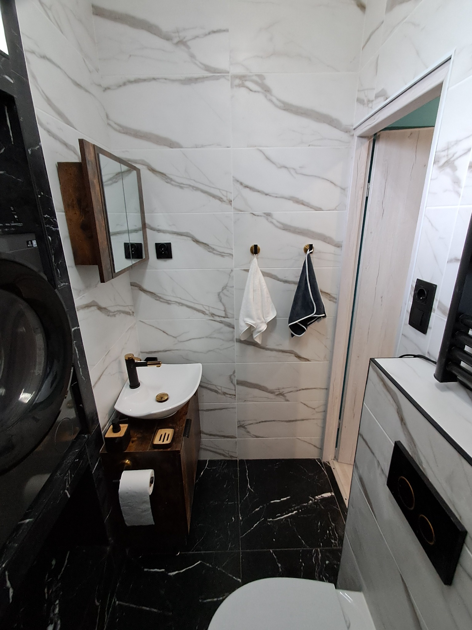 Nowoczesna łazienka z marmurowymi płytkami, czarną armaturą i drewnianymi akcentami. Widoczna umywalka nablatowa, lustro i detale wykończeniowe.