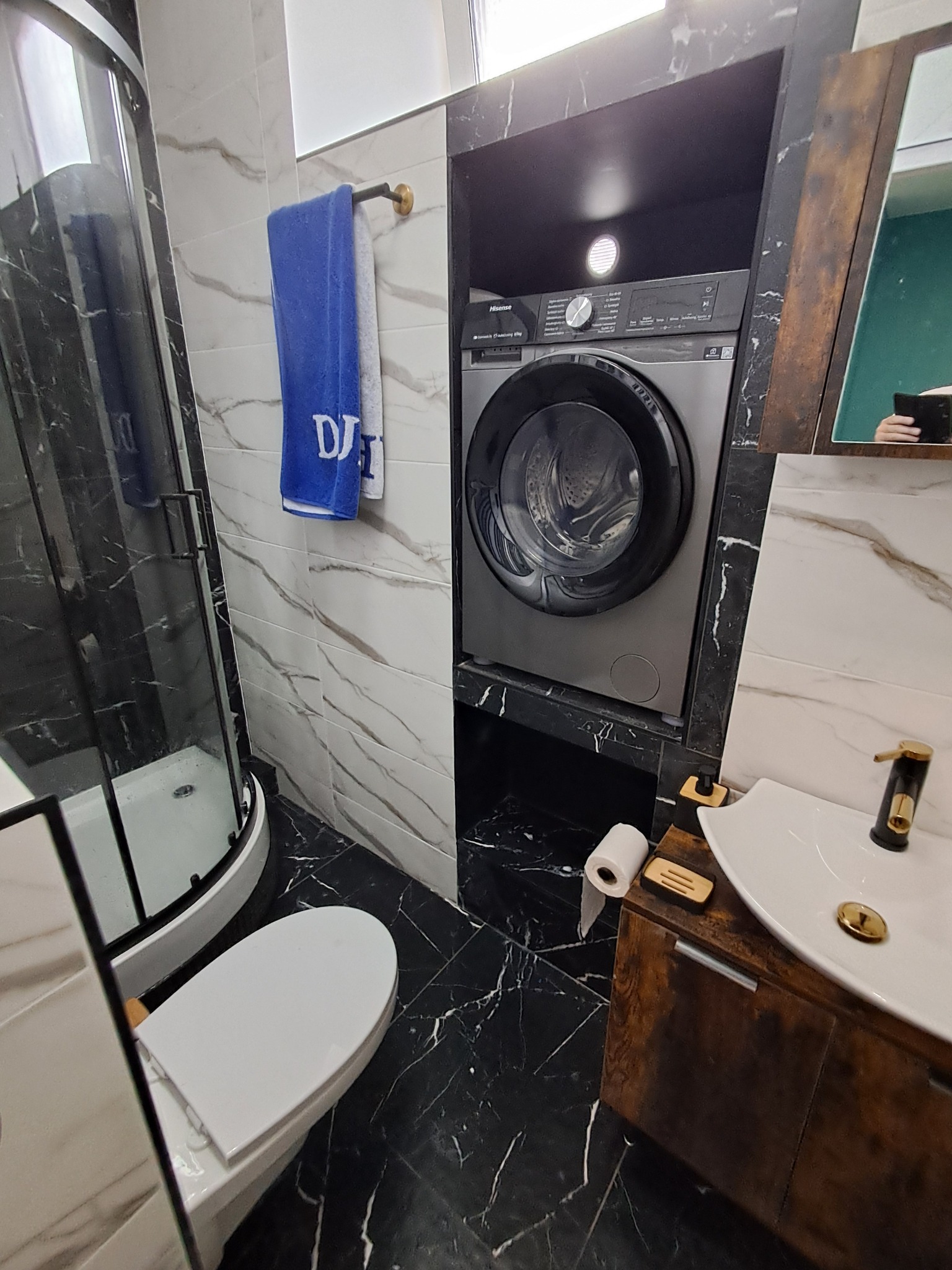 Nowoczesna łazienka w czarno-białej tonacji z marmurowymi akcentami, kabiną prysznicową, toaletą, umywalką z szafką i pralką w zabudowie.